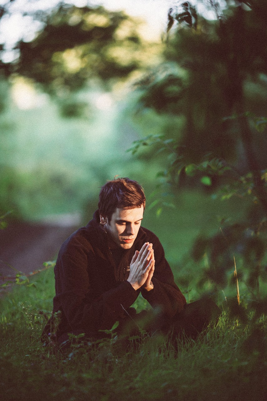 мъж е седнал в тревата в молитвена поза със сключени длани сред природата, дървета, зеленина, вглъбеност, молитва, духовна интелигентност