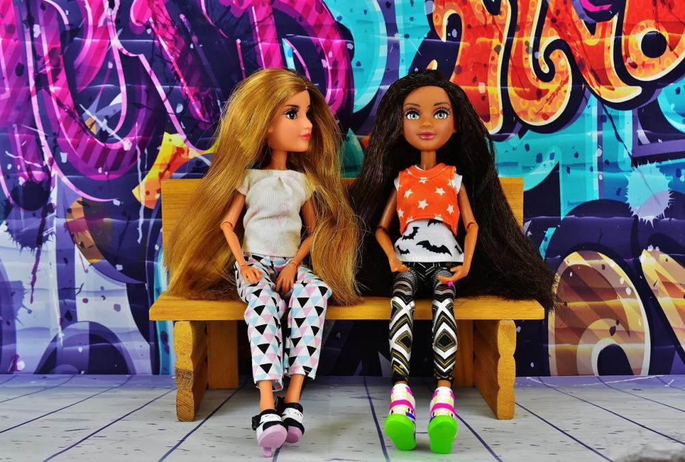 на фона на градски графити в ярки виолетови, сини и оранжеви тонове, две кукли Барби са седнали на пейка и разговарят